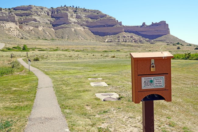 A pet pick up dispenser is seen to the right of a paved trail with a sandstone bluff in the background.