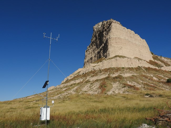 A weather stations sits in a field with a view of a sandstone bluff behind it.
