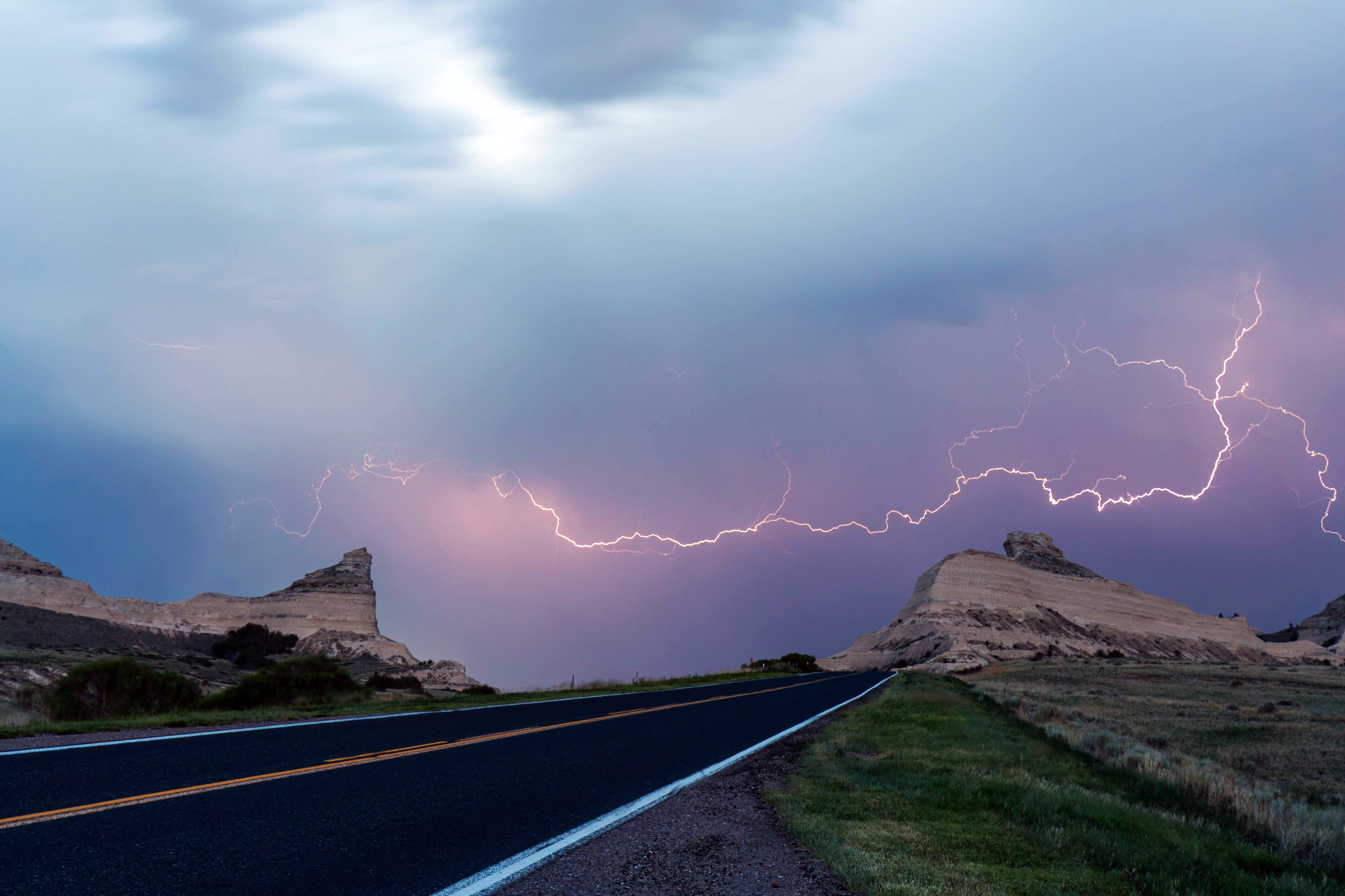 A distant bolt of lightning streaks across the sky over a road between sandstone bluffs.