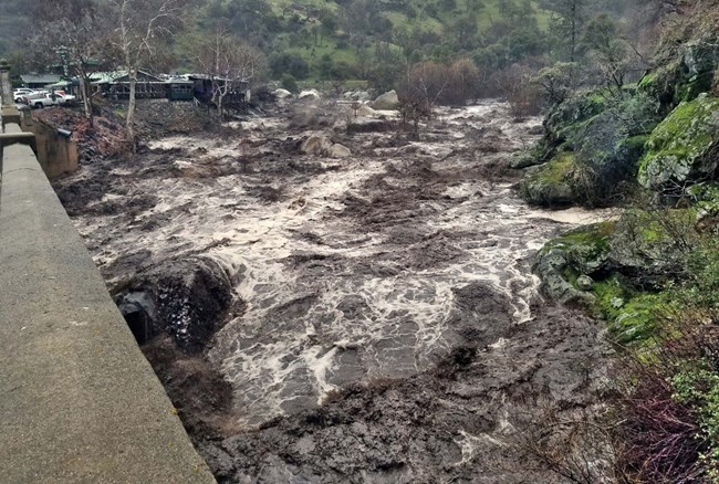 A dark, muddy river full of debris surges down stream and underneath a bridge.