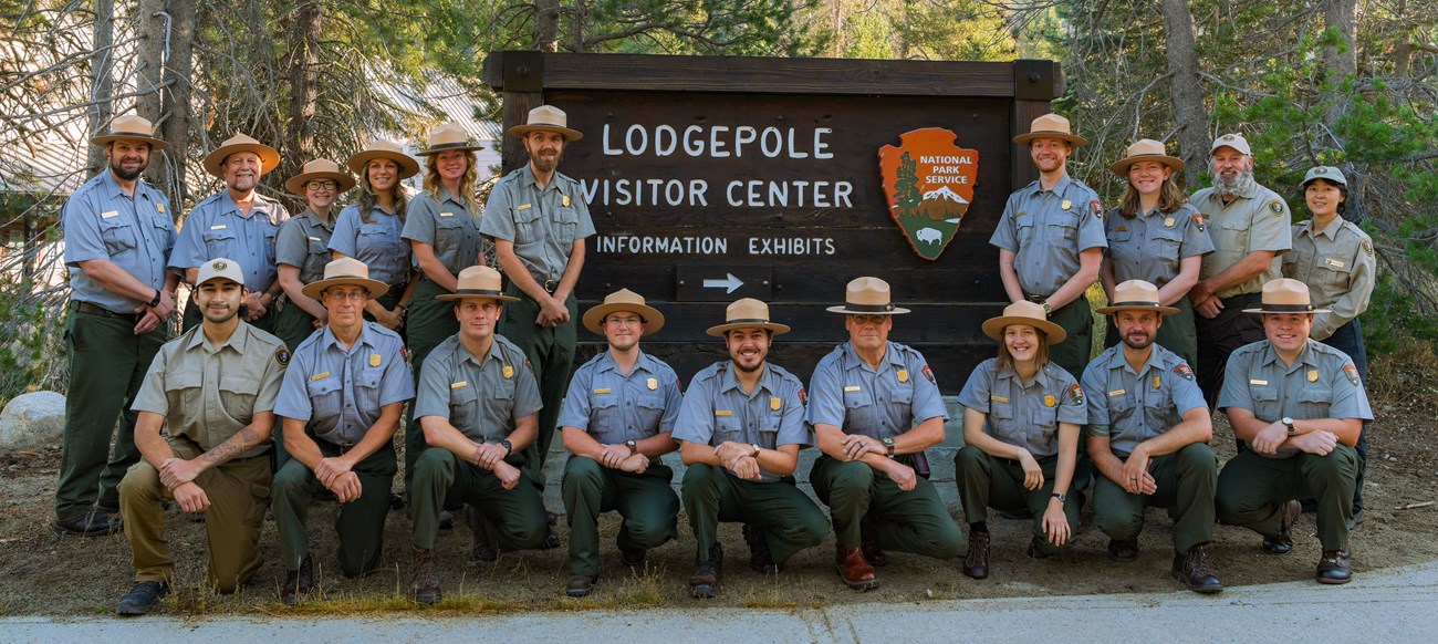 A set of 20 rangers pose in front of the Lodgepole Visitor Center welcome sign. They are smiling and posing for a picture.
