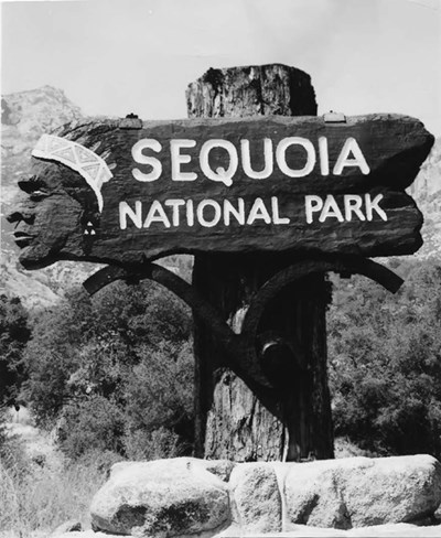 A black and white photograph of Sequoia National Park's historic entrance sign. The sign has an Indian head on the left with the words Sequoia National Park on the right.