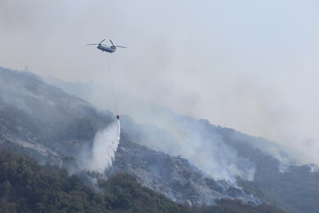 Water drops out of helicopter amidst smoke and active burning of a wildfire below in shrub and oak vegetation.
