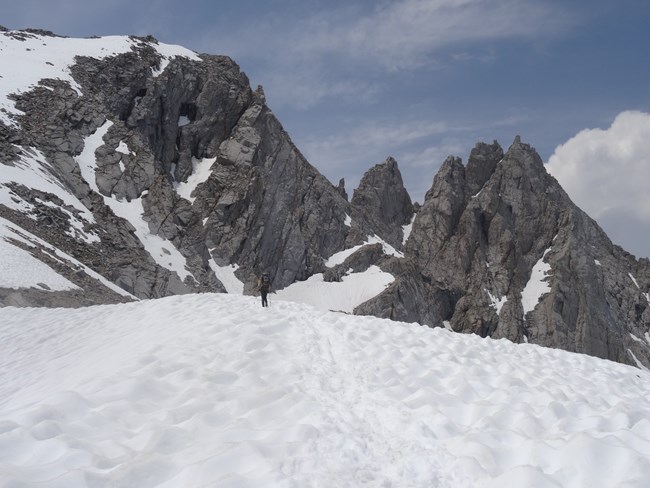 Person on snowfield hiking toward rugged granite peaks.
