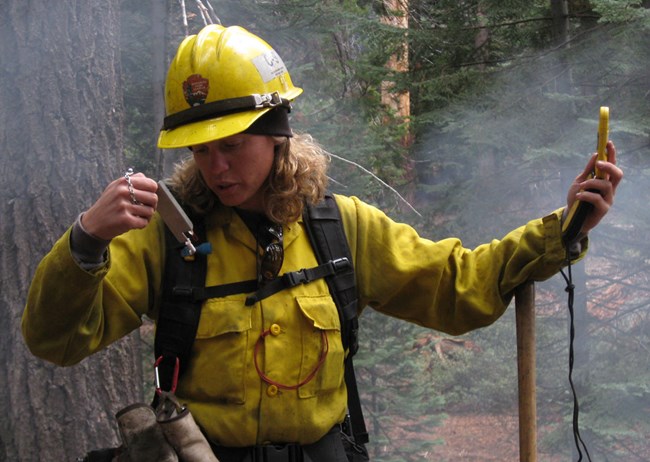 Fire Monitor wearing nomex fire shirt and hard hat examines handheld instrument to read relative humidity near a fire.