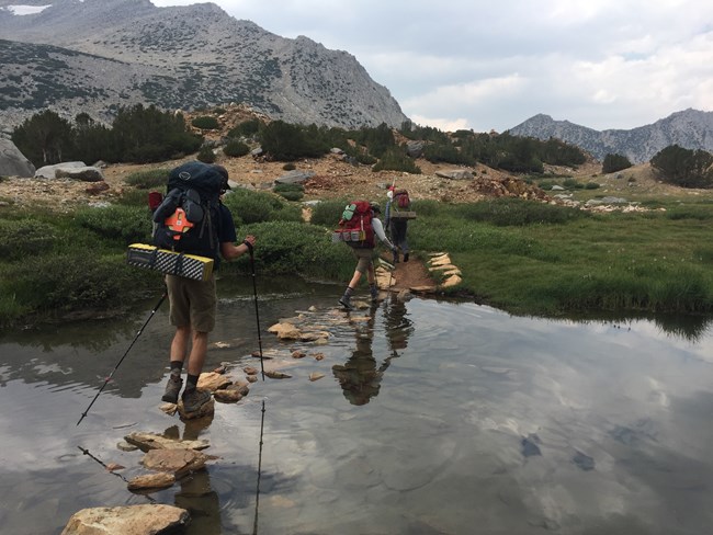 A group of 3 people with backpacks cross a stream by stepping on rocks.