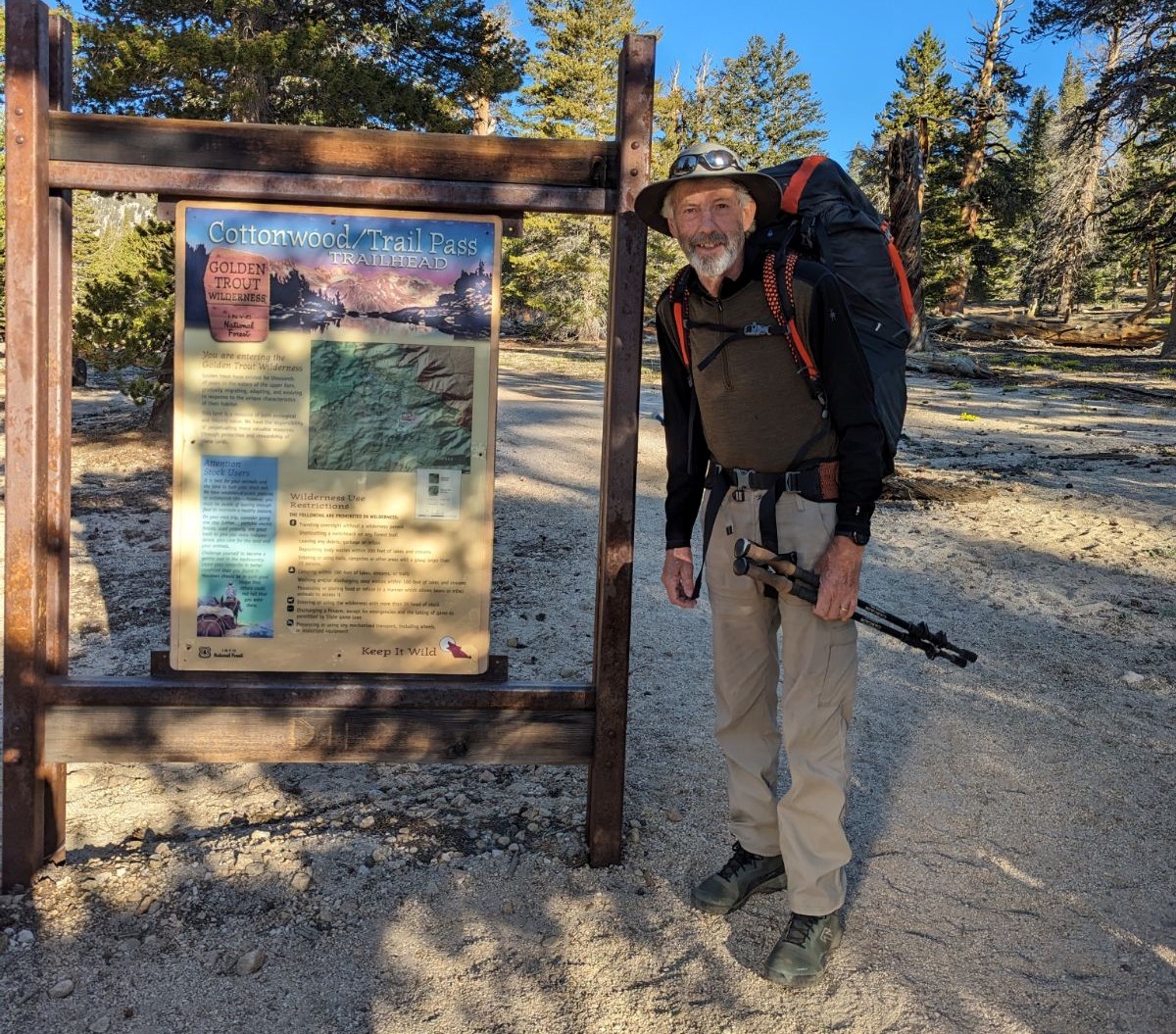 Slim man with gray beard in backpacking gear poses next to a sign reading "Cottonwood/Trail Pass"