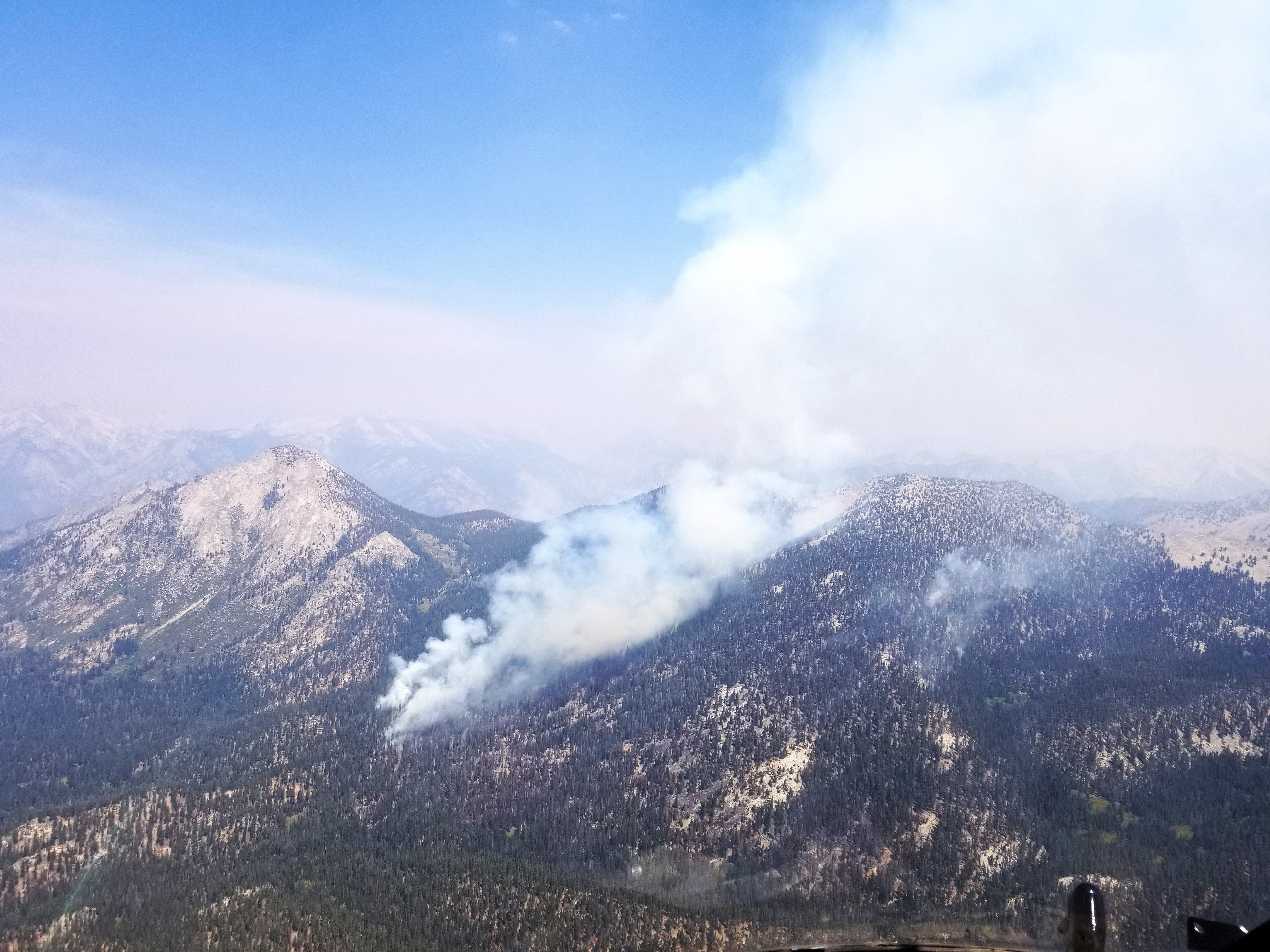 The 2020 Moraine Fire observed from the parks' helicopter shows the fire burning across steep and rugged terrain in designated wilderness. White smoke is seen rising from a forested mountain peak in the middle of the photo.
