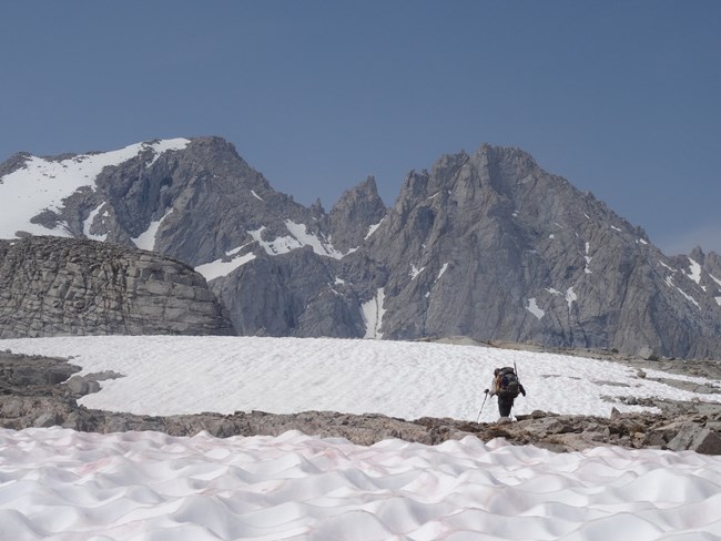 Backpacker stands on rocks overlooking a snowfield and a grand expanse of jagged, rocky mountains.