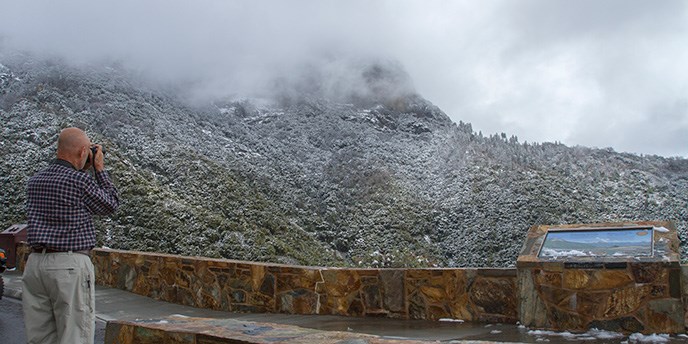 A man taking a photograph of a snow covered hillside from a roadside viewpoint.