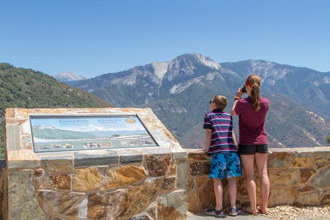 Two visitors at a park overlook gaze out at the views of mountains.