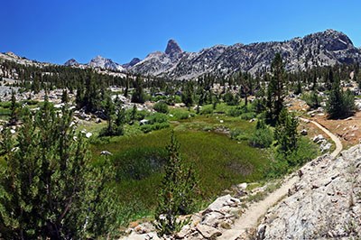 A trail leads past an alpine meadow surrounded by granite mountains