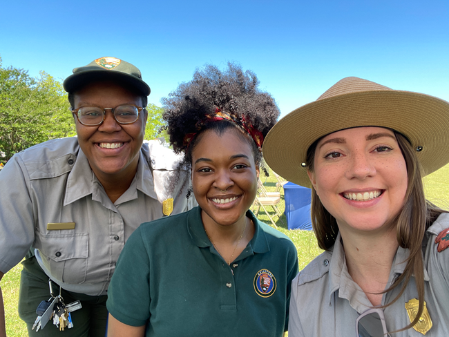 A trio of smiling young women, two are black and one is white. Two are wearing a park service uniform, one has a green volunteer polo.