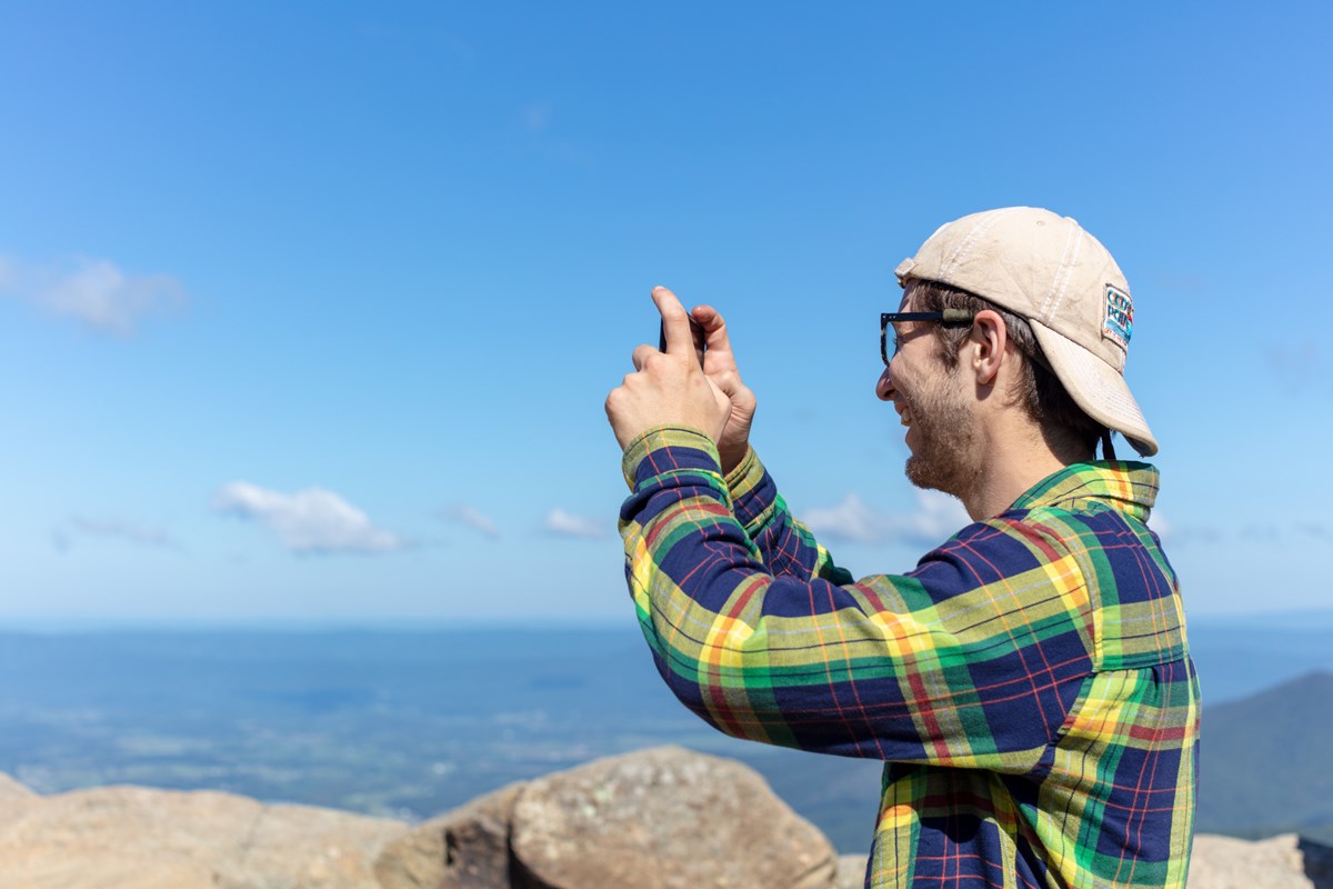 A young man stands on top of a scenic overlook and takes a picture with his phone.