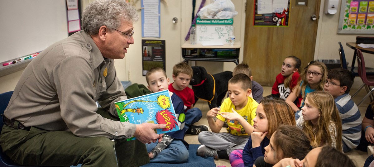 A male park ranger reads a book to a group of children sitting at his feet in a classroom.