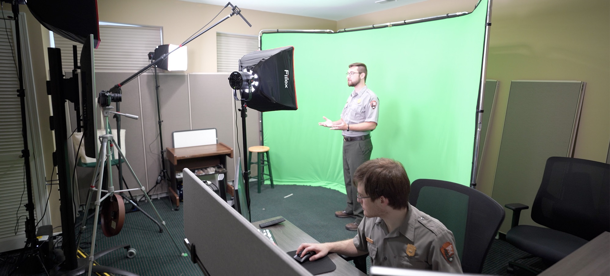 A behind the scenes look of a studio set up with a green screen. One ranger stands in front of the green screen while another runs a soundboard.