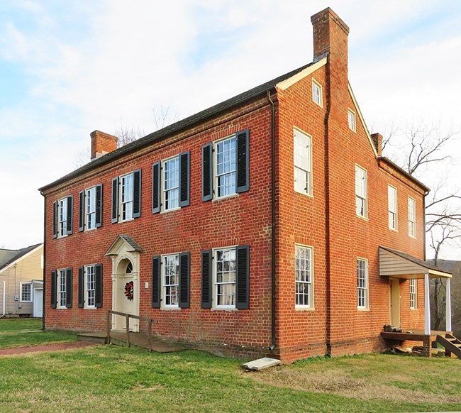 A red brick house from an angle, with windows along the first and second floor, featuring black shutters, a beige front door with a green wreath and a red brick walkway leading to the front door