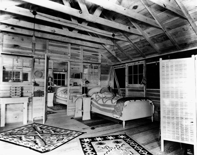 Interior of a log cabin showing rustic wood furnishings and woven rugs.