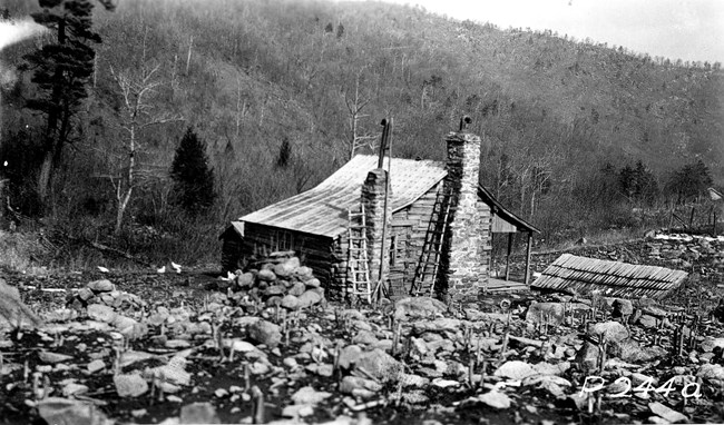 A black and white photo of a log cabin with stone chimneys, surrounded by drystone walls and set against a forested hillside.