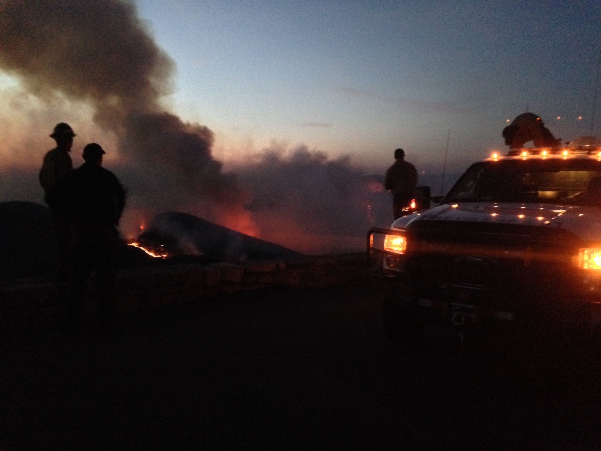 Firefighters at night observing an orange glow and smoke rising below them from an overlook
