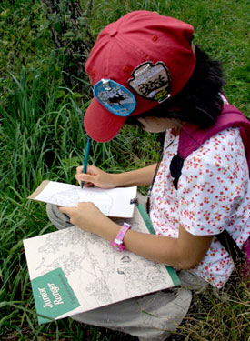 A young visitor wearing a red Junior Ranger hat and patches, sits in the grass of Big Meadows working on her Junior Ranger book. A young visitor wearing a red Junior Ranger hat and patches, sits in the grass of Big Meadows working on her Junior Ranger book.