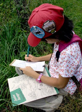 A young visitor wearing a red Junior Ranger hat and patches, sits in the grass of Big Meadows working on her Junior Ranger book.