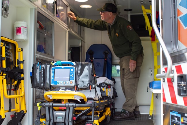 ambulance interior with stretcher, EKG machine, and PSAR ranger reaching for medical supplies.
