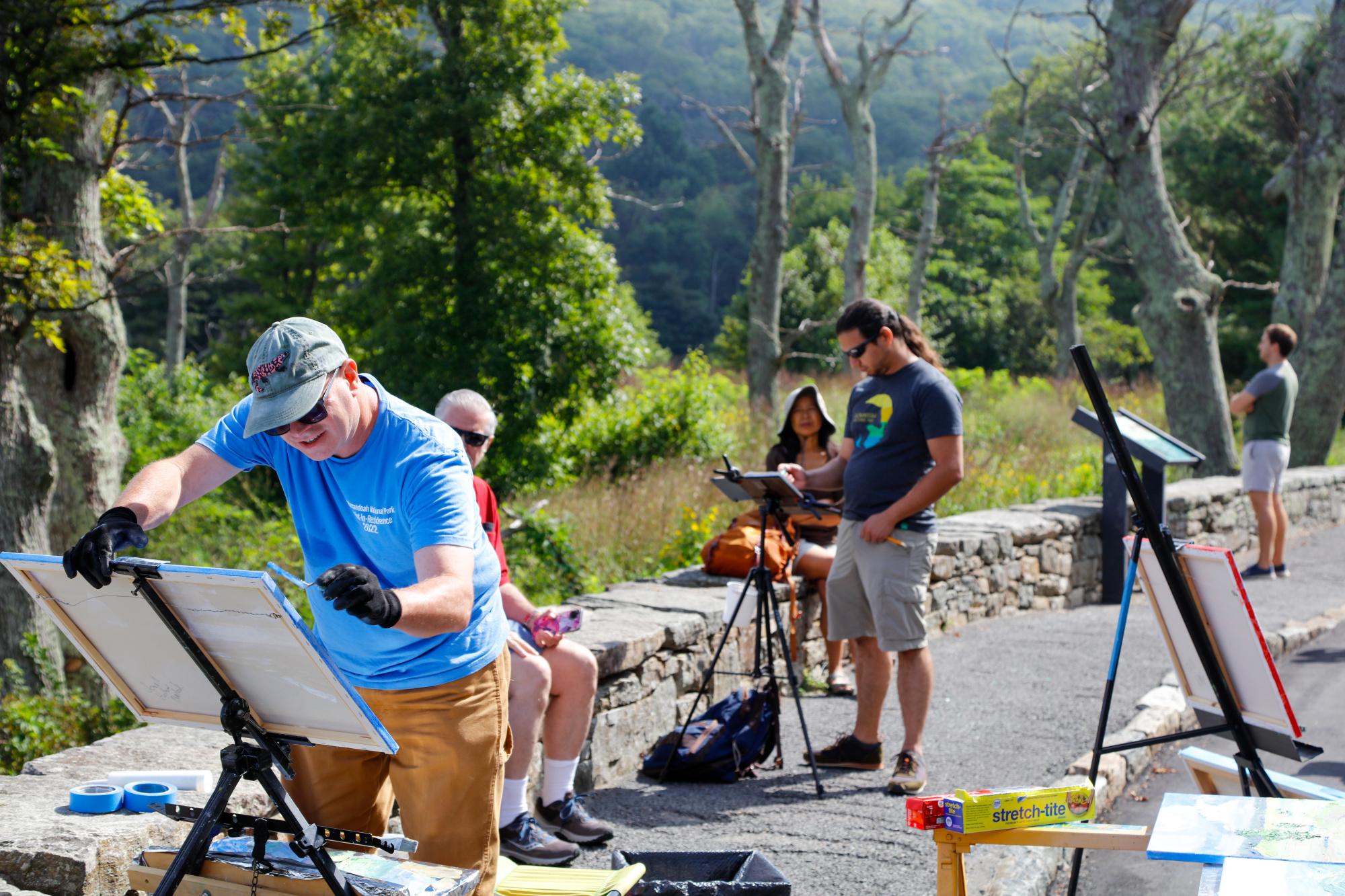 An artist giving a plein air painting demonstration surrounded by art supplies and visitors.
