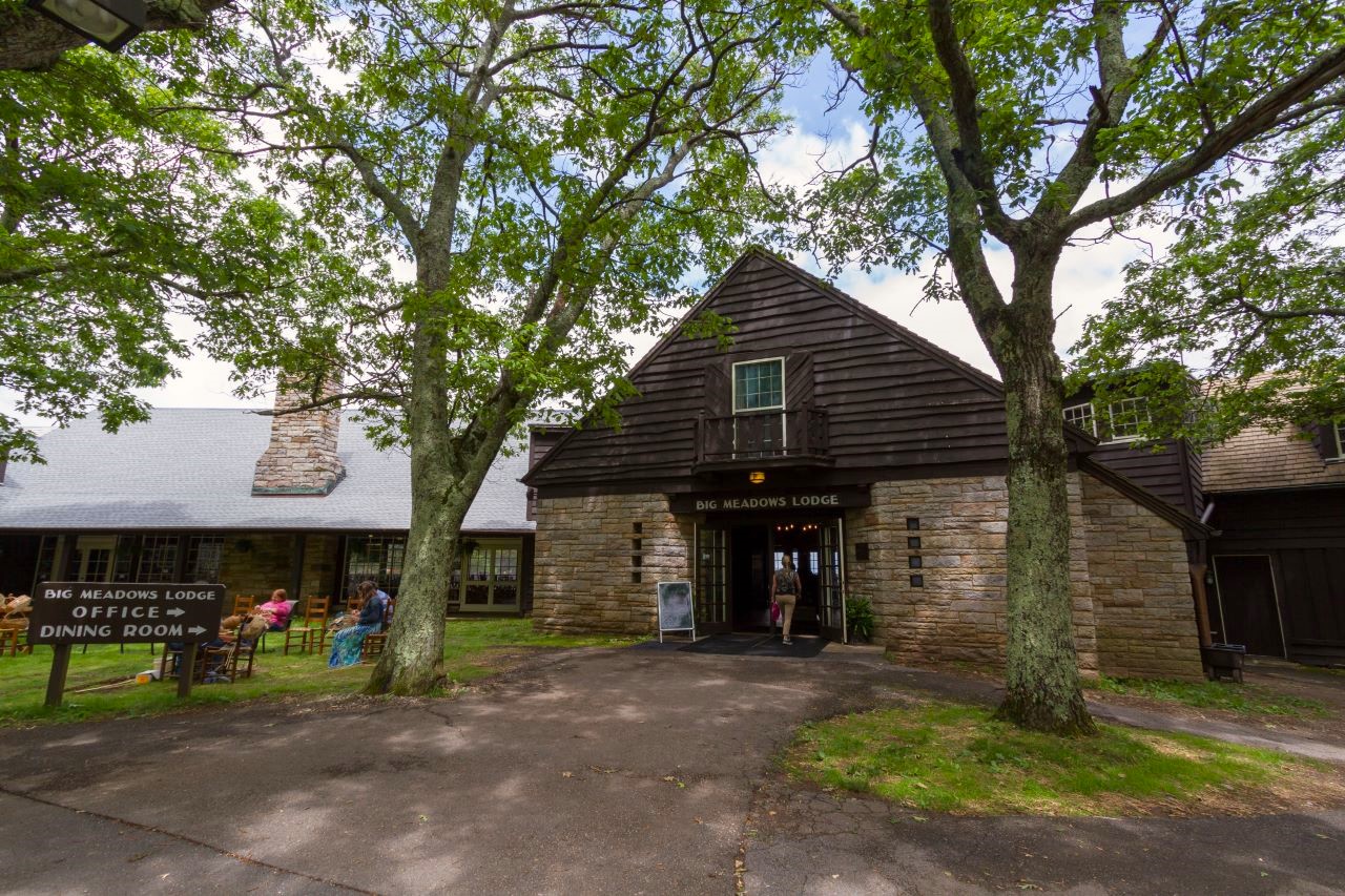 Big Meadows Lodge A brown stone and dark brown sided building labeled "Big Meadows Lodge" stands in the center. A woman walks through a doorway in the center of the building. A window above the doorway. In front of the building are two large trees and green grass on either