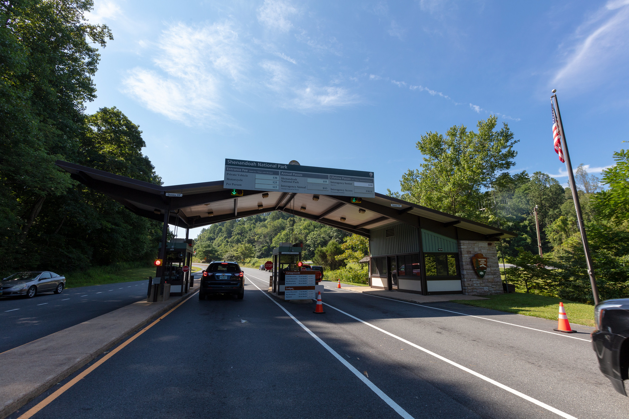 Swift Run Entrance Station A National Park Service entrance station building with two lanes for driving through. A car is stopped under the building at a station. A car exits the park on a road to the left. A U.S. Flag is on a flag pole at right.