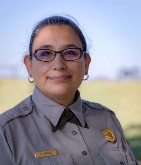 A woman with brown hair in a tight bun. She is wearing a gray ranger shirt with NPS badge, green cross tie, and oval glasses.
