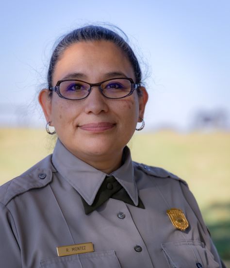 Raquel Montez A woman with brown hair in a tight bun. She is wearing a gray ranger shirt with NPS badge, green cross tie, and oval glasses.