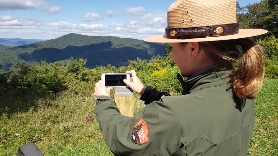 A female park ranger holds a phone on a wooden post to take a picture of the viewshed in front of her.