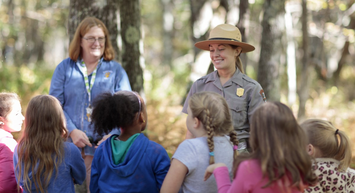 A group of children listen to a female Park ranger talk in the middle of the woods.