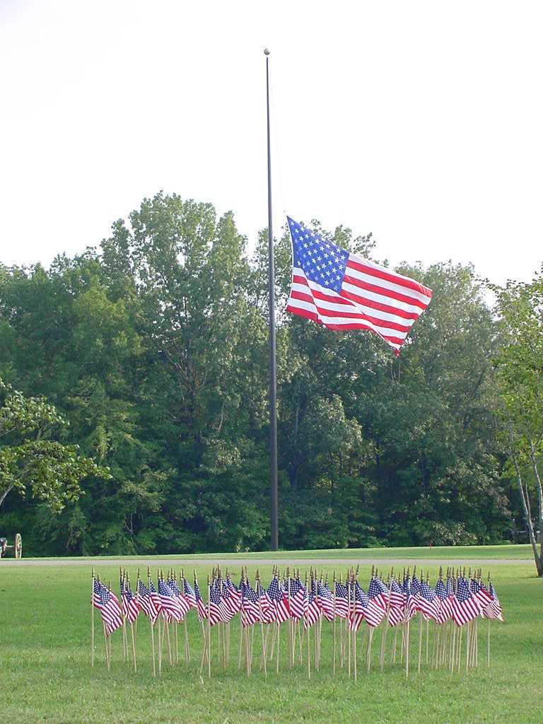 A large U.S. flag in background flying at half staff with several smaller flags in foreground