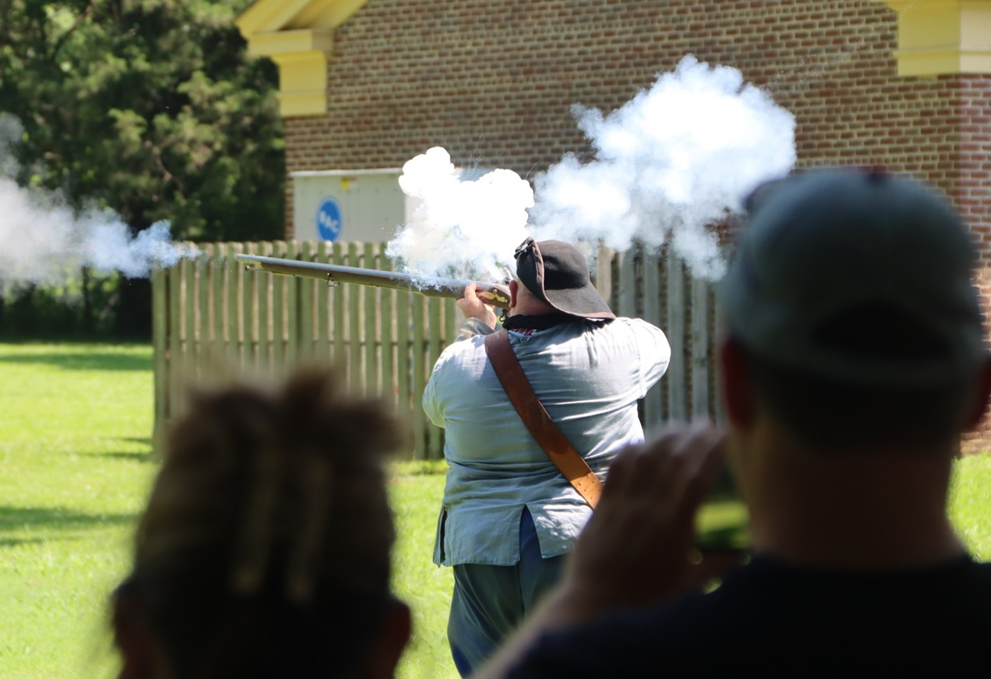 a person in revolutionary war clothing fires a musket with silhouettes of two people watching in the foreground