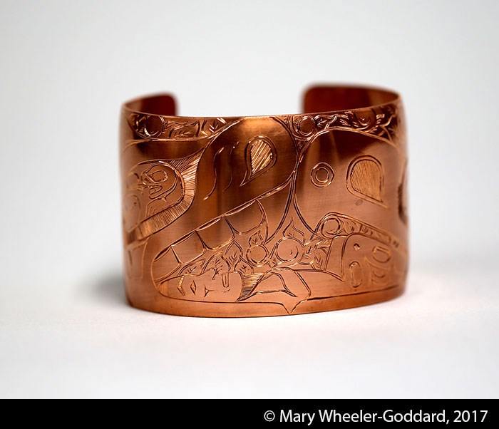 A wide copper cuff bracelet with formline design carvings on the surface.