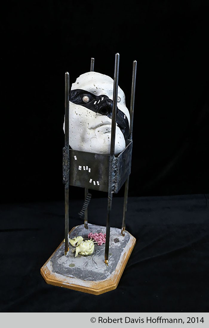 Sculpture made of a white plaster mask with a black stripe crossing over the left eye down to the right side of the chin, supported by four metal bars with metal plates attached between them. Beneath this are pieces of broken dolls. Sculpture made of a white plaster mask with a black stripe crossing over the left eye down to the right side of the chin, supported by four metal bars with metal plates attached between them. Beneath this are pieces of broken dolls.