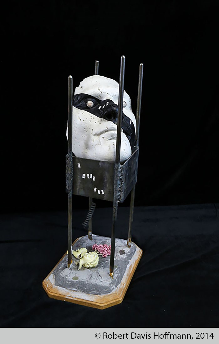 Sculpture made of a white plaster mask with a black stripe crossing over the left eye down to the right side of the chin, supported by four metal bars with metal plates attached between them. Beneath this are pieces of broken dolls.