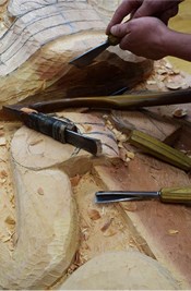 Wood being carved with a chisel, with two adzes resting nearby.