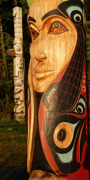 Close-up of the Mother Earth figure on the Centennial Pole with bright red, black and blue paint on her salmon/hair, and a faded totem pole standing in the back left.