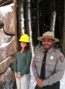 Park Ranger in uniform and volunteer wearing a yellow hardhat stand alongside a totem pole lightly covered in snow.