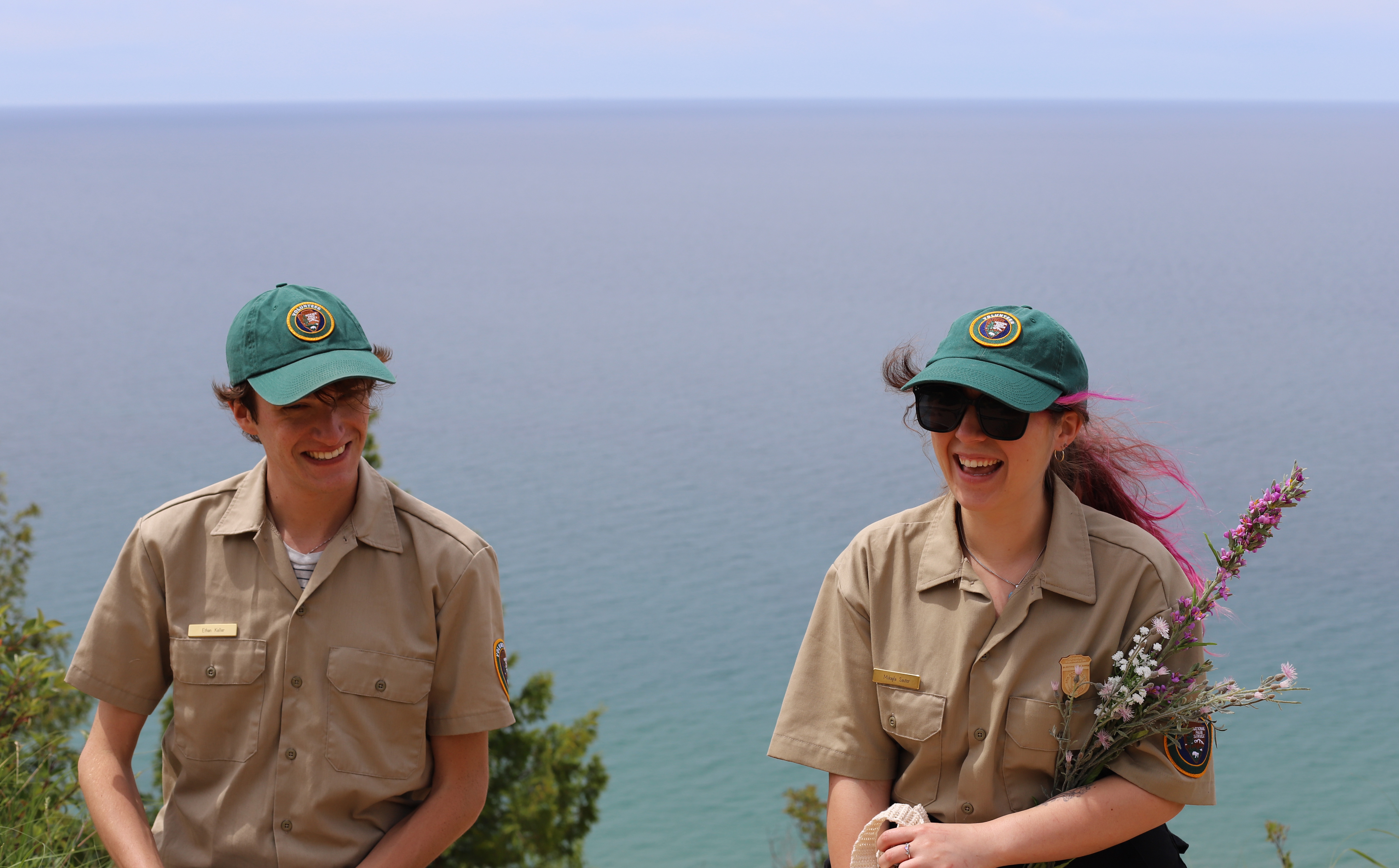 Two interns wearing volunteer hats smile in front of Lake Michigan.