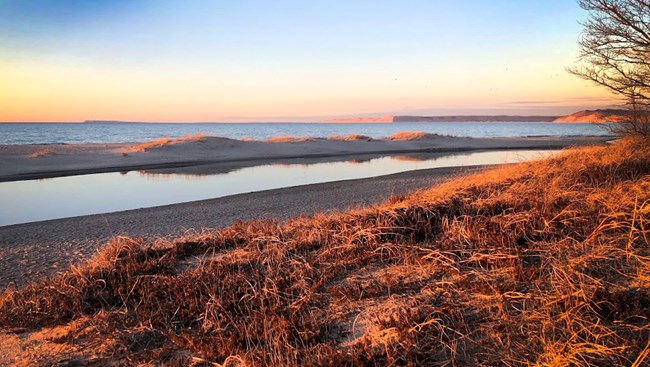 A river flows out into a large lake with beach and dune grass surrounding.