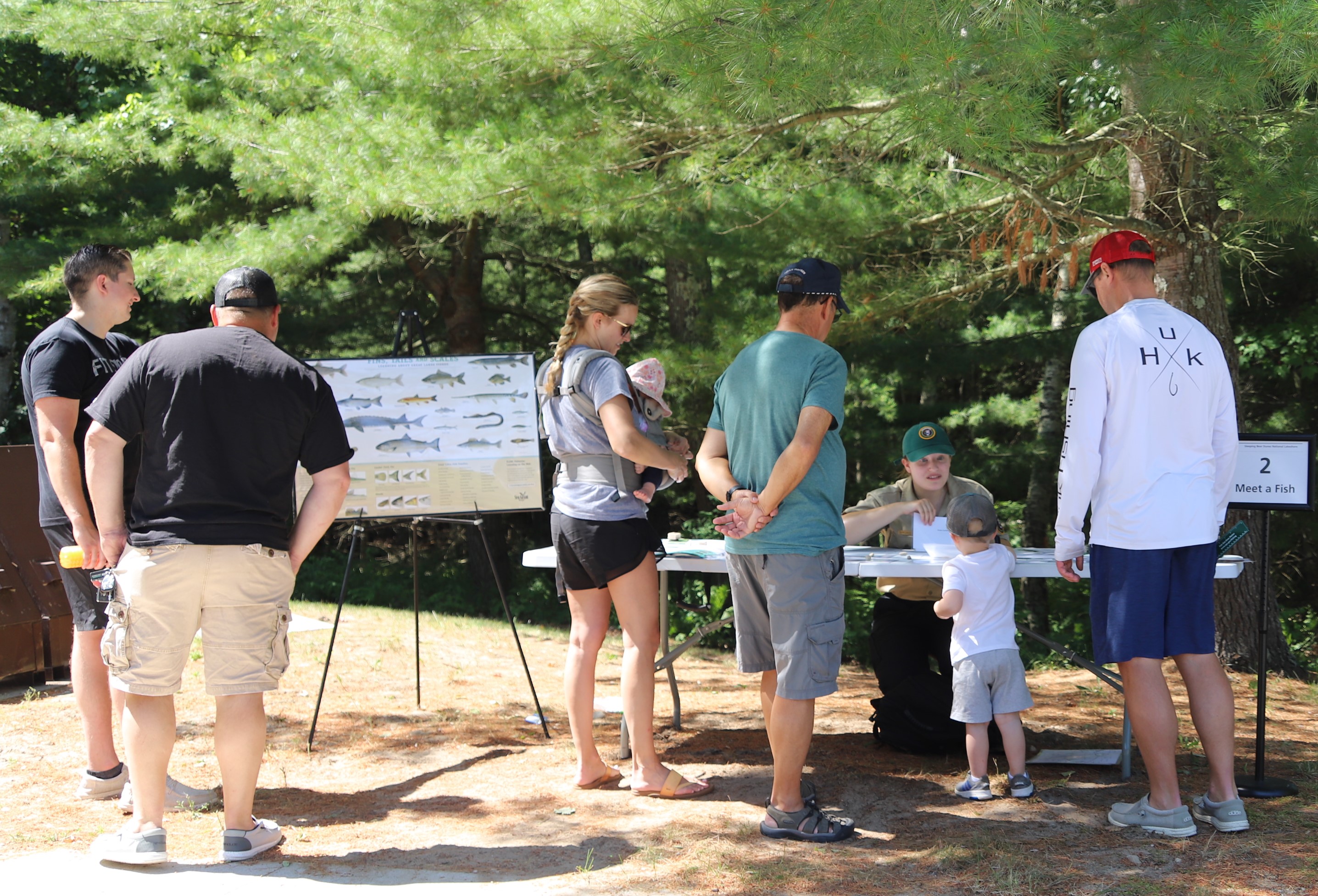 Jr_Angler_Day.jpg A small child stands next to a table surrounded by adults. A volunteers kneels on the other side of the table holding a picture of a fish.