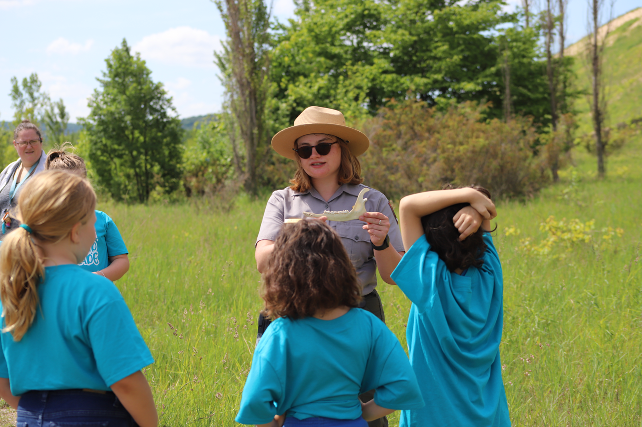 A ranger wearing a flat straw hat and sunglasses displays a deer jawbone to a group of elementary school students in matching blue shirts