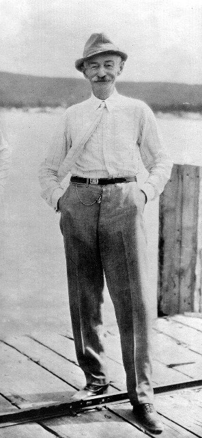 Black and white image of a smiling older man standing on a wooden dock. He is wearing a hat, light long sleeved shirt and tie, and pants with a belt and pocket watch chain.