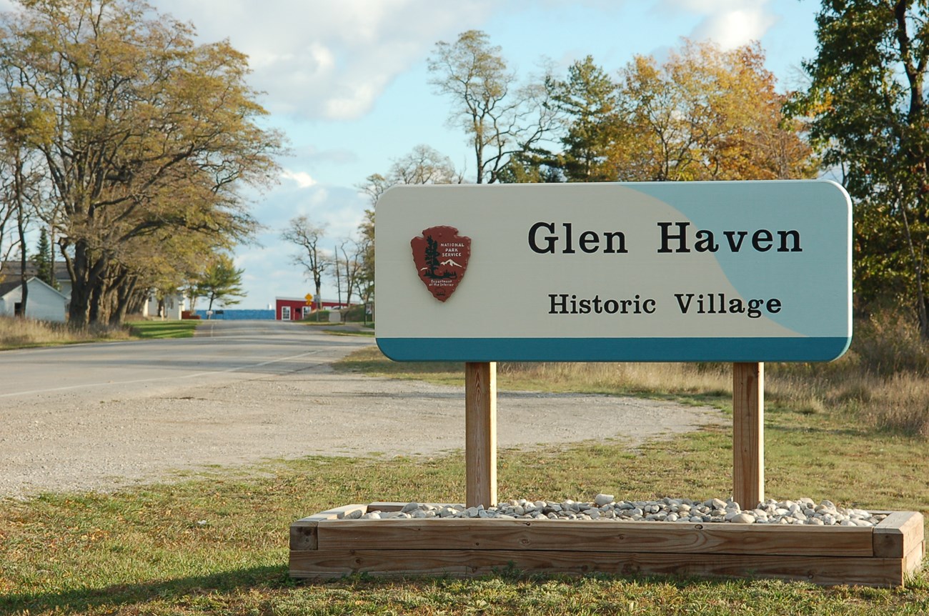 A sign with an NPS arrowhead symbol stands in front of a road lined with trees. The sign says "Glen Haven, Historic Village".