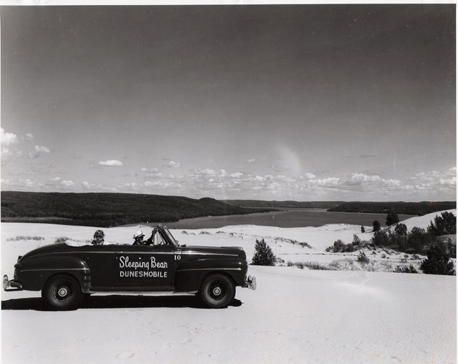 A black and white picture of a car driving on a dune, with a lake and forested hills in the background. Text on the car says "Sleeping Bear Dunesmobile".