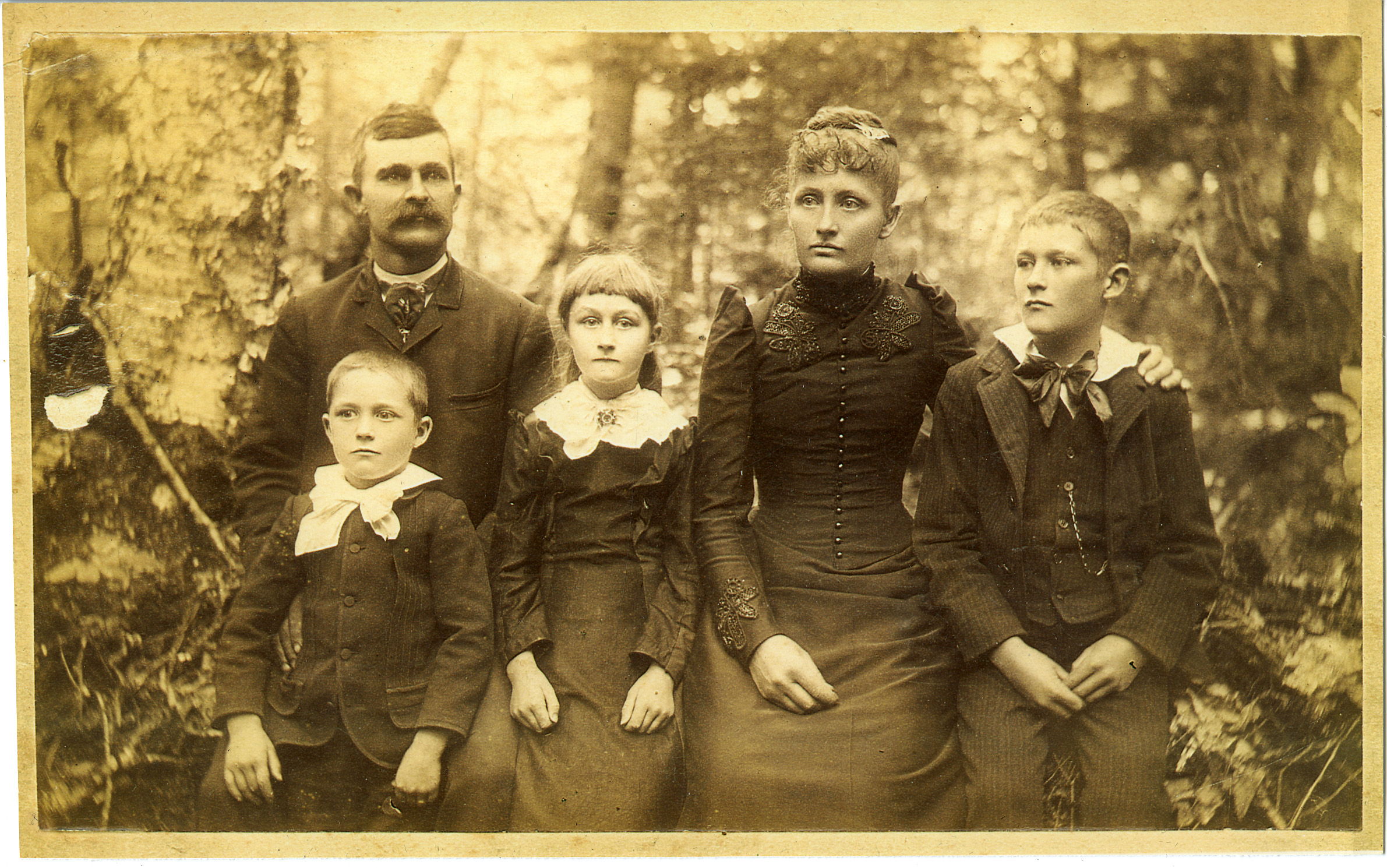An aged black and white family photograph taken in a wooded setting. There are two parents and three children. One boy is sitting on his father's lap.